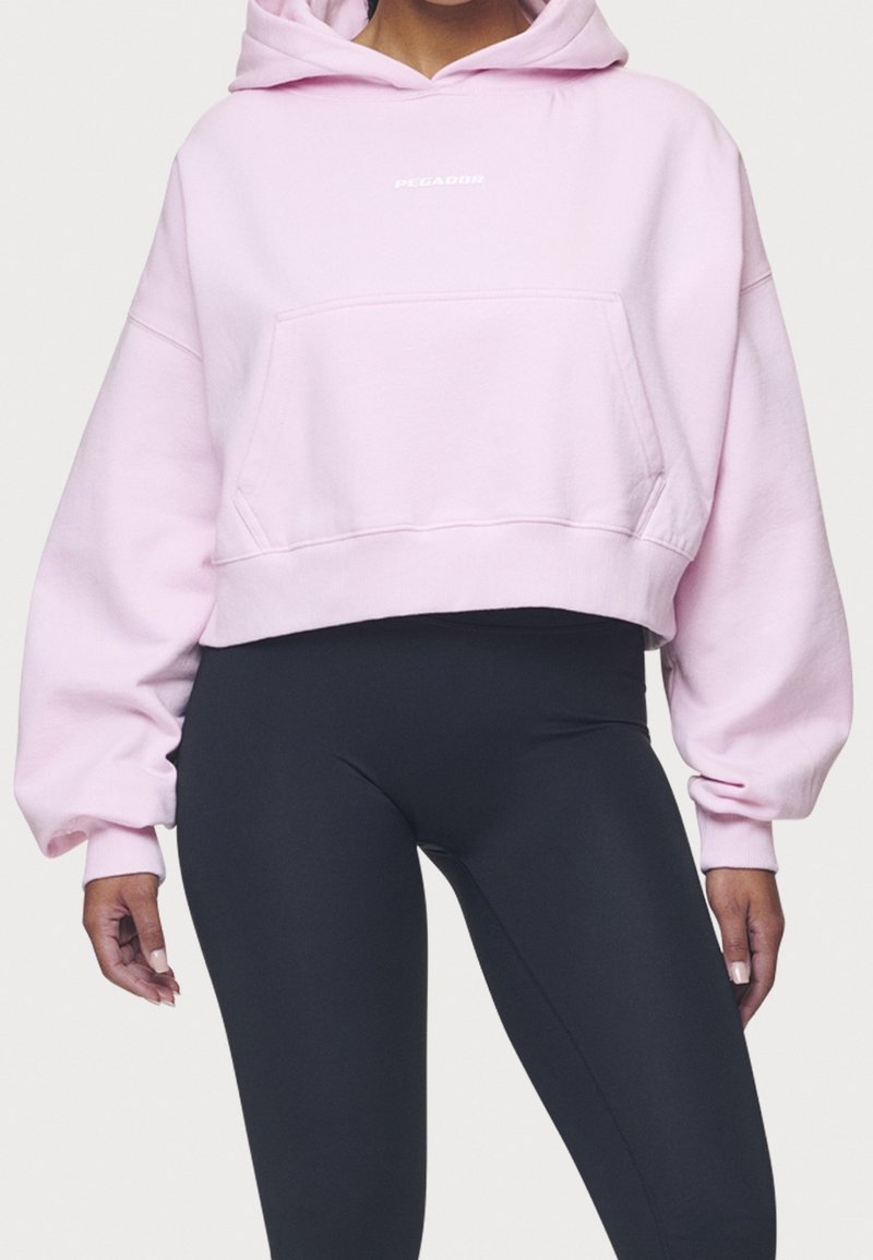 Cropped pink hoodie with a front pocket and logo, featuring wide sleeves and a soft texture. Paired with black leggings.