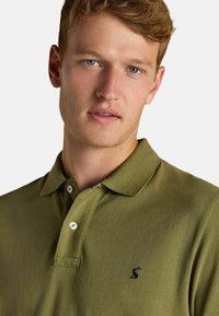 Olivgrünes Polohemd aus strukturiertem Stoff, mit einem Kragen und drei Knöpfen, sowie einem kleinen navyfarbenen Logo auf der Brust.