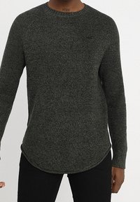 Mörkgrön stickad tröja med rund halsringning, långa ärmar och böjd nederkant. Har ett texturerat mönster och diskret logotypdetalj.
