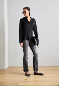 Schwarze langärmlige Bluse mit Knöpfen, graue Schlagjeans, schwarze Ballerinas mit Schleifen, hält eine schwarze Clutch, steht auf einem Holzboden.