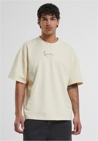 Kremo spalvos trumpomis rankovėmis marškinėliai, pagaminti iš medvilnės, su apvaliu kaklu ir rudu logotipo dizainu priekyje. Lygus audinys, laisva forma.