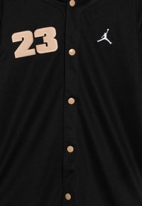 Camiseta deportiva negra con el número "23" en beige cosido en el lado izquierdo del pecho. Presenta un logo blanco, cierre de botones y tela texturizada.