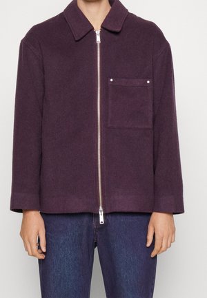 Man wearing a dark purple zip-up jacket with a collar and a single chest pocket, paired with blue jeans.