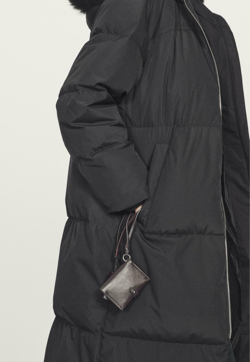 Schwarze Daunenjacke mit glatter Textur, ausgestattet mit einem silbernen Reißverschluss und seitlichen Taschen. Eine kleine, dunkelbraune Lederbörse hängt an einem Riemen.