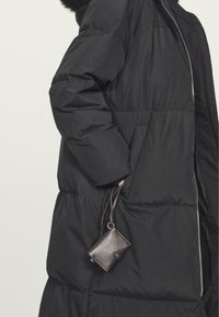 Zwarte pufferjas met een gladde textuur, voorzien van een zilveren rits en zijzakken. Een kleine, donkerbruine leren portemonnee hangt aan een lus.