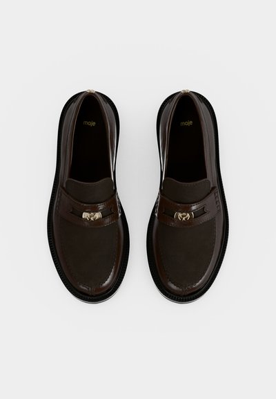 Ζευγάρι σκούρα καφέ δερμάτινα loafers με στρογγυλεμένες μύτες και λεπτομέρειες με χρυσή αγκράφα, εμφανιζόμενα από πάνω σε απλό φωτεινό φόντο.