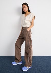 Top beige cortado com decote em V, calças largas castanhas com riscas brancas laterais e sapatilhas atleticas azuis com detalhes brancos.