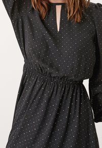 Černé šaty s malými béžovými puntíky, výstřih s kulatým otvorem, dlouhé rukávy a nařasený elastický pas, které nosí osoba s hnědými vlasy.