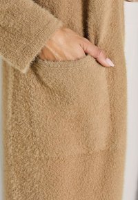 Hand mit manikürierten Nägeln, die in die flauschige, camelbraune Pullover-Tasche gleitet und das weiche, strukturierte Strickgewebe sowie das Ärmel-Detail zeigt.