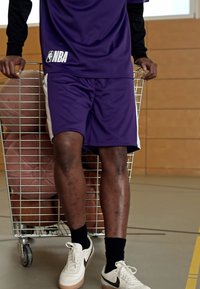 Pantaloni da basket a rete viola con accenti bianchi, indossati con calze nere e scarpe da ginnastica bianche, in piedi accanto a un carrello di metallo con palloni da basket.