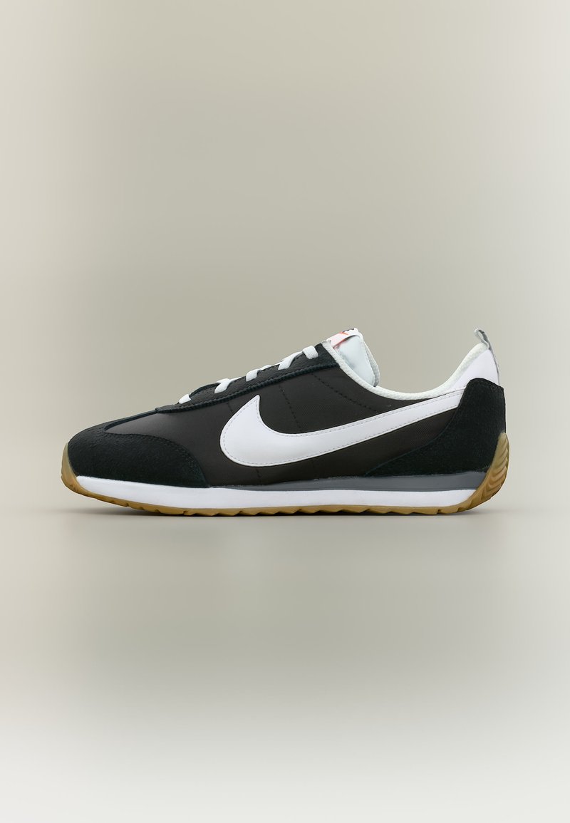Μαύρο και λευκό αθλητικό παπούτσι Nike με καουτσούκ σόλα, λευκό λογότυπο Swoosh, δέσιμο με κορδόνια και σχεδιασμό χαμηλού παπουτσιού σε ουδέτερο φόντο.