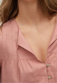 Světle růžová lněná blůza s výstřihem do V, vertikálními sklady a dvěma zelenými knoflíky vpředu. Jemná textura a uvolněný střih.