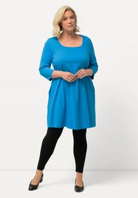 Blaues knielanges Tunika-Kleid mit Dreiviertelärmeln, quadratischem Ausschnitt, seitlichen Taschen und kombiniert mit schwarzen Leggings und Absatzschuhen.