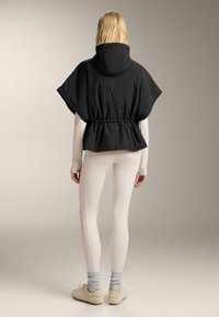 Zwarte poncho met capuchon en een getailleerde taille, gecombineerd met witte legging. De poncho heeft een gladde textuur en een ontspannen ontwerp voor comfort.