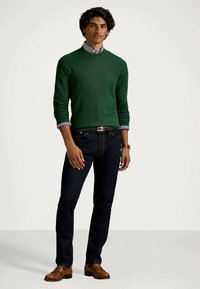 Man standing wearing green sweater, striped collared shirt, dark jeans, brown belt, watch, and brown leather shoes on plain background.