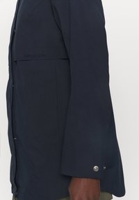 Marineblauer wasserdichter Mantel mit lässigem Schnitt, ausgestattet mit einem Knopfverschluss und einem silbernen Druckknopf am Ärmel. Gesticktes Logodetail am Ärmel.