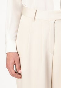 Pantalons taille haute de couleur crème avec des plis, fabriqués à partir d'un tissu texturé. Comprend une large ceinture et des détails de poches latérales.
