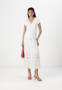 White short-sleeve top with lace neckline, paired with a white ankle-length skirt featuring eyelet details and button front, and a red bag.