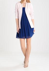 Růžová přiléhavá sako přes tmavě modré šaty ke kolenům s nařasenou strukturou. Šaty mají rozšířenou sukni a průhledné detaily. Béžové podpatky.