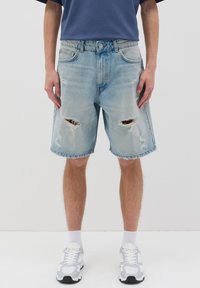 Pantalones cortos de mezclilla azul claro con bordes desgastados y parches rasgados en las rodillas. Lleva una camiseta azul marino y zapatillas blancas.