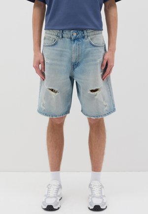 Bermuda jeans blu chiaro con orli sfrangiati e toppe strappate alle ginocchia. Indossati con una t-shirt blu navy e sneakers bianche.