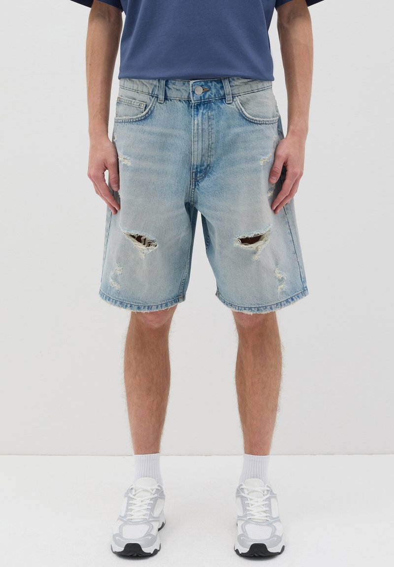Pantalones cortos de mezclilla azul claro con bordes desgastados y parches rasgados en las rodillas. Lleva una camiseta azul marino y zapatillas blancas.