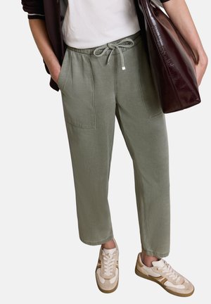 Osoba nosząca luźne oliwkowozielone spodnie ze sznurkiem, białe trampki i niosąca na ramieniu dużą ciemnobrązową skórzaną torbę.