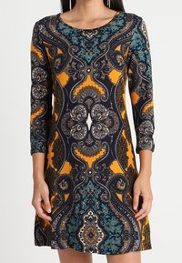 Robe imprimée arborant un motif paisley en bleu marine, orange et bleu sarcelle. Elle possède un encolure ronde et des manches trois-quarts. Tissu doux.