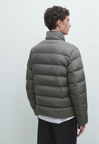 Graue Pufferjacke mit hohem Kragen, gestepptem Design und elastischen Bündchen, getragen über einem weißen Hemd und schwarzen Hosen. Glattes, strukturiertes Gewebe.