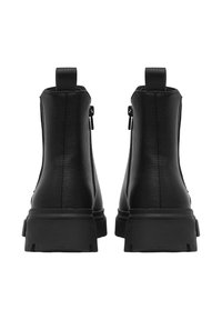 Un paio di stivaletti neri in pelle con suole in gomma spessa e cerniere laterali, visti da dietro su uno sfondo bianco.