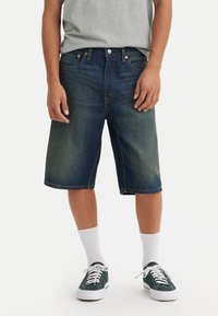Tummansiniset farkkuhousut, joissa on haalistuneita alueita, suora leikkuu ja käännetty helma. Yhdistetty valkoisiin urheil socksisiin ja vihreisiin kangastossuihin.