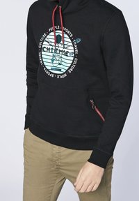 Svart sweatshirt med grafiskt tryck i mitten, röda dragsnören och en sidoficka med dragkedja. Har ribbade mudd och nederkant.