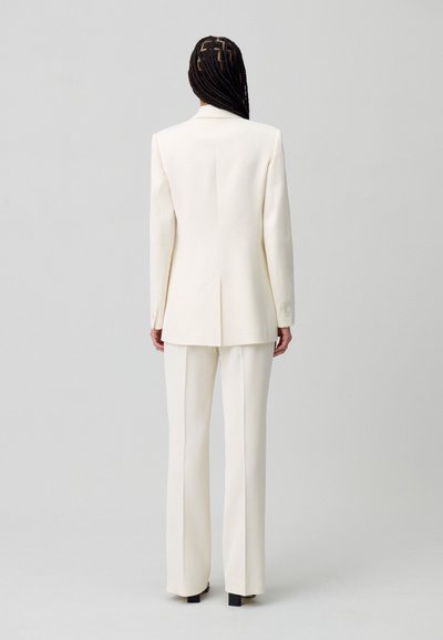 Traje blanco a medida con una chaqueta larga y pantalones rectos. Textura suave, diseño minimalista y silueta estructurada.