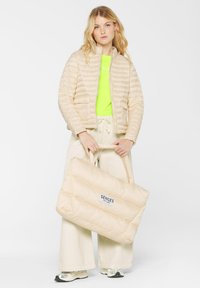 Beige Daunenjacke mit horizontalen Nähten, hohem Kragen und zwei Fronttaschen. Das Modell hält eine passende gesteppte Tasche mit der Aufschrift "SENSES".