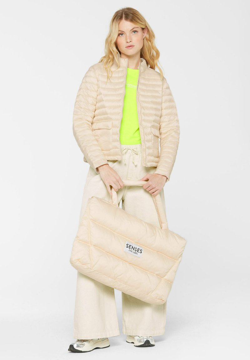 Beige Daunenjacke mit horizontalen Nähten, hohem Kragen und zwei Fronttaschen. Das Modell hält eine passende gesteppte Tasche mit der Aufschrift "SENSES".