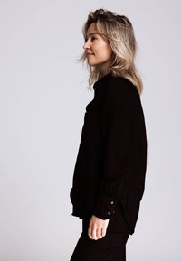 Schwarzes Cord-Hemd mit entspanntem Schnitt, vorne mit Tasche, Knopfleiste und Manschetten mit silbernen Knöpfen. Vertikal gerippte Textur.