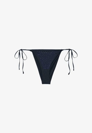 Kobieta z mokrymi włosami w czarnym bikini z wiązaniem po bokach i czarnych klapkach, stojąca na tle jednolitego brązowego tła.