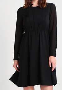 Svart klänning med långa ärmar, plisserad framdetalj, hög halsringning och utsvängd kjol. Tillverkad av ett mjukt tyg med ett flödande fall.