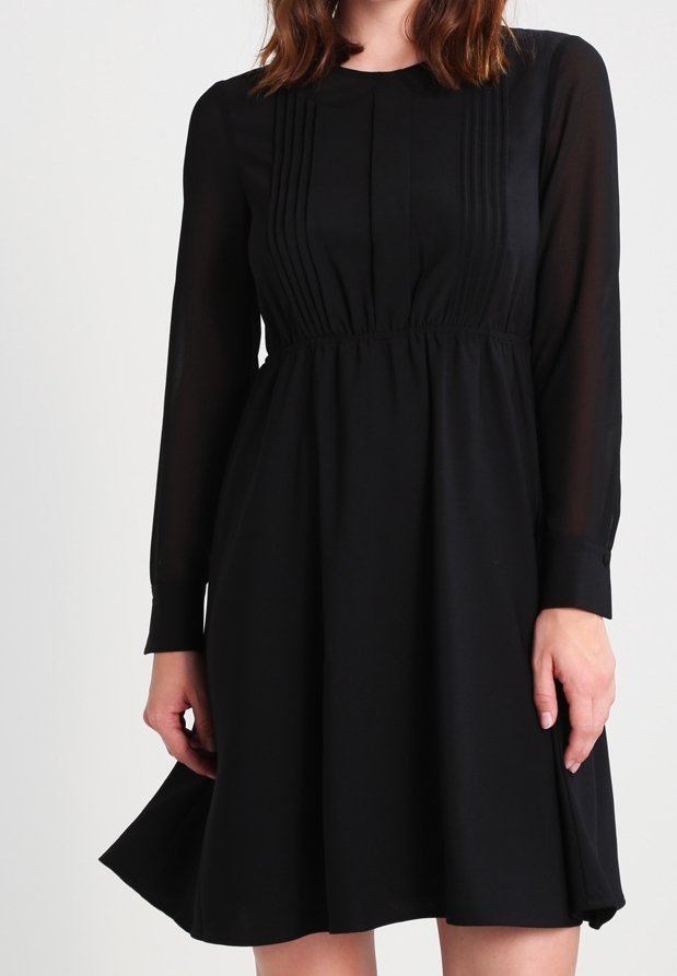 Svart klänning med långa ärmar, plisserad framdetalj, hög halsringning och utsvängd kjol. Tillverkad av ett mjukt tyg med ett flödande fall.