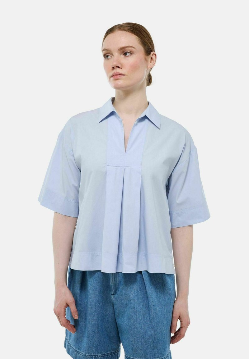 Blouse légère bleu clair à manches courtes avec col, détail de pli sur le devant et texture douce. Associée à un short en denim bleu ample.