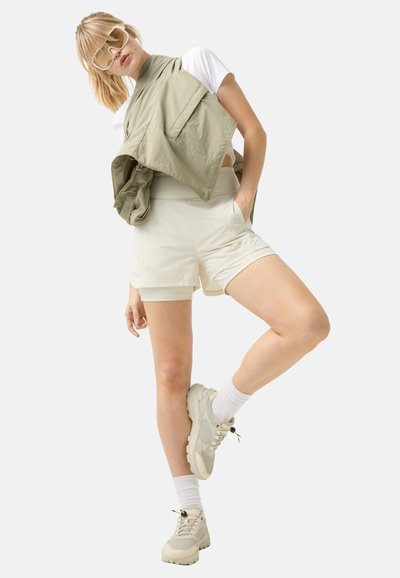 Lichtgroene mouwloze jas over een wit shirt, gecombineerd met crèmekleurige shorts en beige sneakers. Witte sokken accentueren de schoenen.