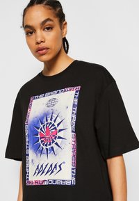 Black cotton t-shirt featuring a large graphic print with blue and pink accents, including a stylized logo and text elements. Short sleeves.