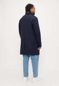 Cappotto in lana blu navy con taglio classico, spacco posteriore singolo e colletto sagomato, abbinato a jeans azzurri e sneaker bianche.
