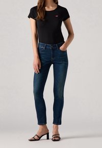 Top nero a maniche corte con un piccolo logo rosso, abbinato a jeans skinny blu scuro e tacchi a strappo neri. Tessuto morbido; vestibilità slim.