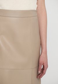 Beige bőrszoknya sima textúrával és látható varrásokkal. Magas derék kialakítás, bemutatva egy kézzel, amely a szöveten nyugszik.
