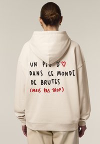 Felpa color crema con vestibilità ampia, caratterizzata da testo nero e rosso sulla schiena. Il testo include un simbolo a forma di cuore e una frase in francese.