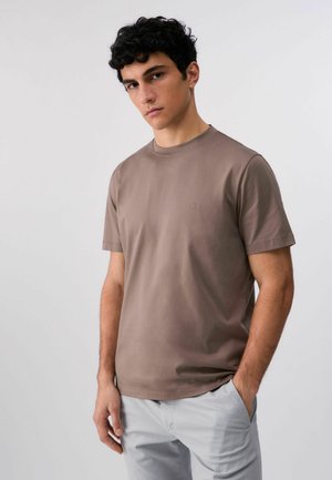 Giovane uomo con capelli scuri e ricci che indossa una maglietta marrone tinta unita a maniche corte e pantaloni grigio chiaro, in piedi davanti a uno sfondo chiaro.