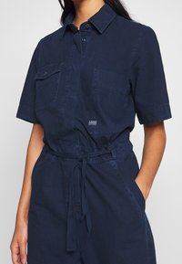 Marinblå kortärmad jumpsuit i bomull, med två bröstfickor, midjeband och knappstängning. Strukturerad tyg.