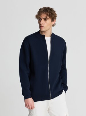Uomo che indossa un maglione blu navy con cerniera sopra una camicia bianca e pantaloni bianchi, guarda a sinistra contro uno sfondo grigio uniforme.