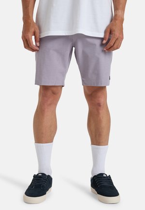 Lilac shorts with a smooth texture, side pockets, and a small black logo tag. Paired with white socks and navy suede shoes.